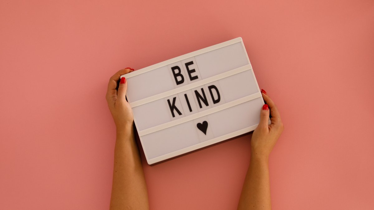 two hands holding a box with letter saying "be kind" with a heard, salmon pink background
