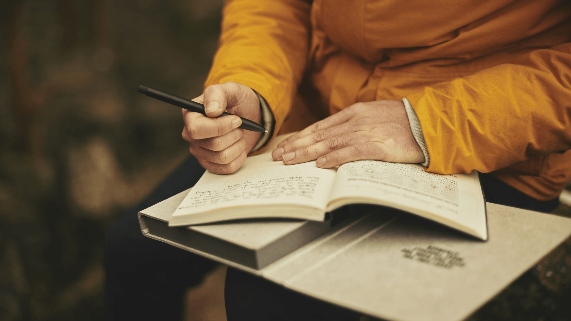 man with orange jacket, pen in the right hand, writing in his journal