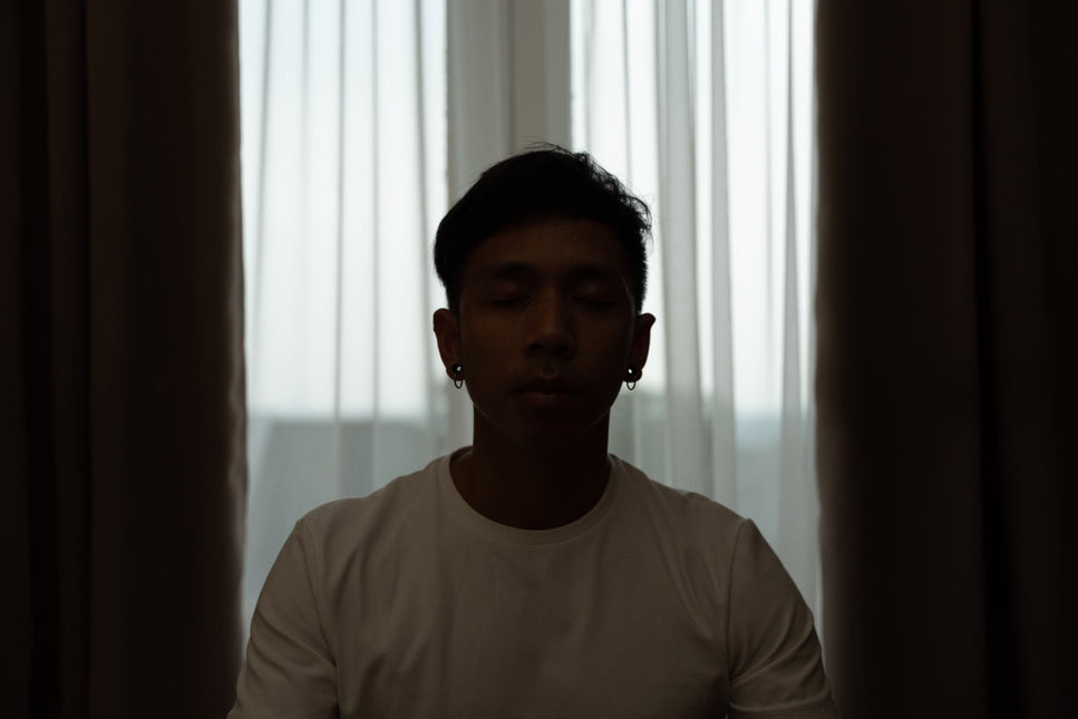 Bayu coach at Life Architekture, wearing white t-shirt, eyes closed, dark room, with curtains in the background